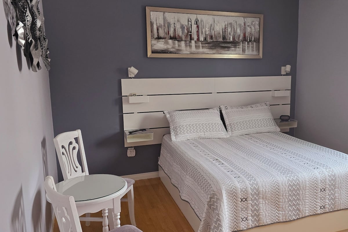 Un dormitorio con una cama, mesitas de noche, una silla, una pequeña mesa, un cuadro en la pared y una ventana.