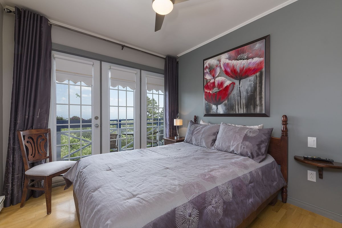 Un dormitorio con una cama grande, una silla, un cuadro y vistas a través de una ventana.