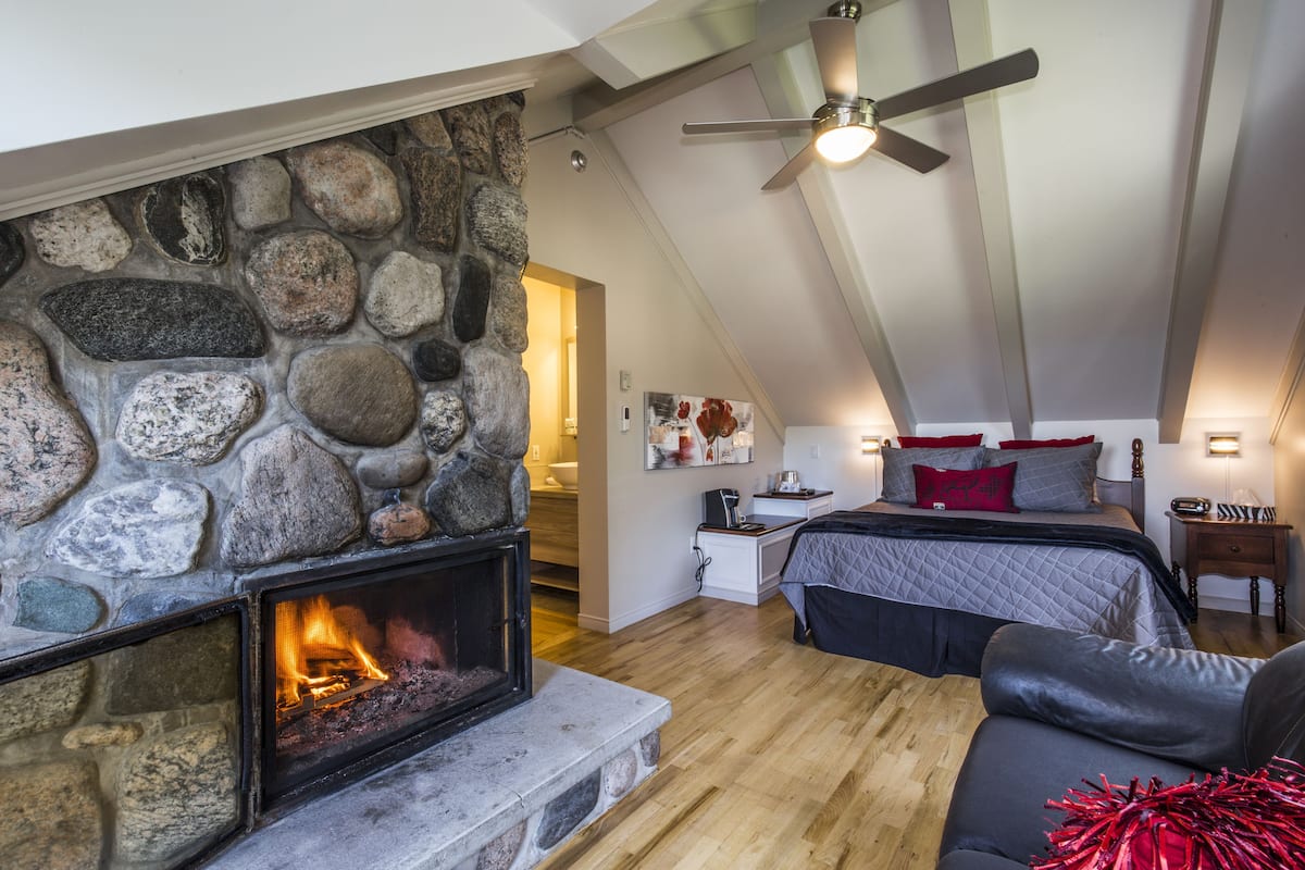 Un dormitorio acogedor con chimenea de piedra, cama con almohadones rojos, mesita de noche y ventilador de techo.