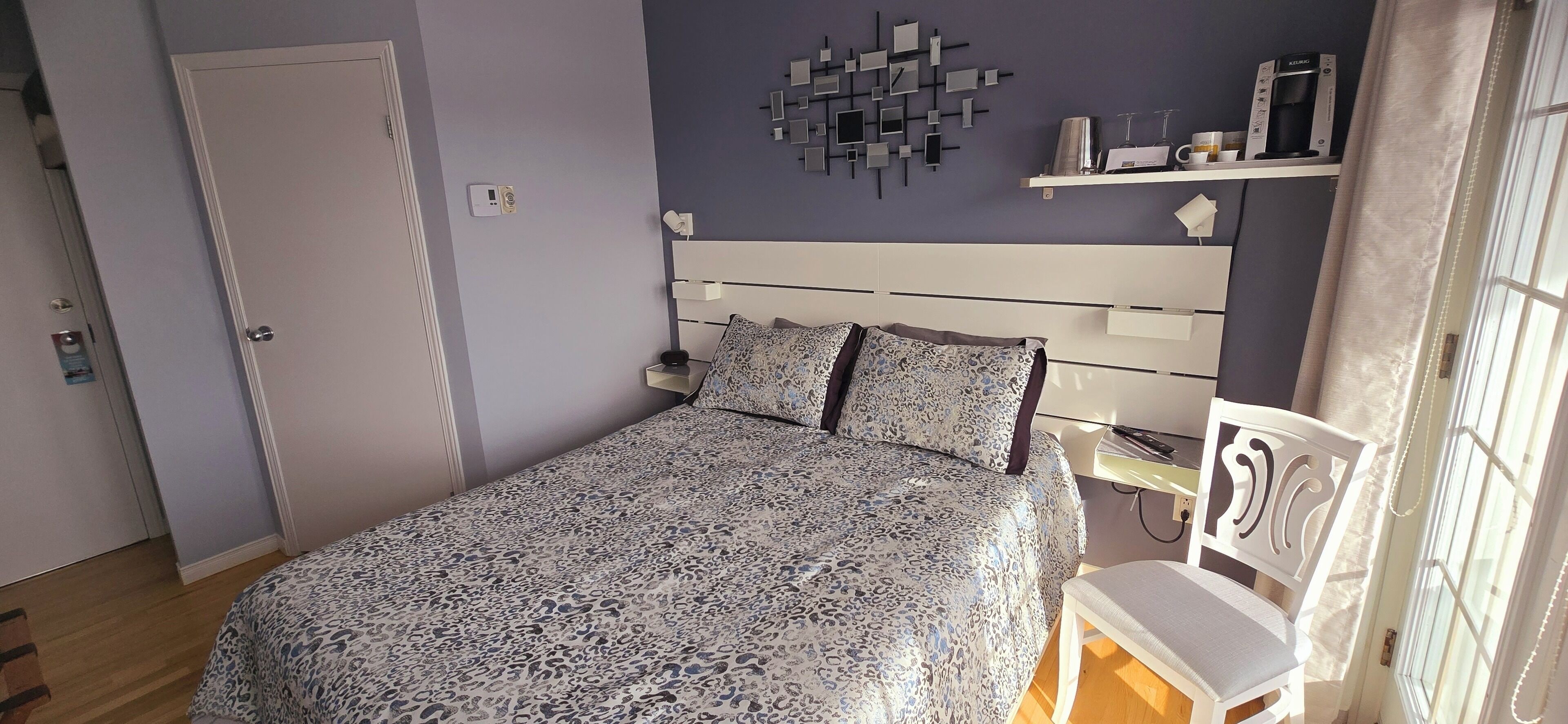 A bedroom with a bed, a nightstand, a chair, a wall-mounted shelf, and a window with curtains.