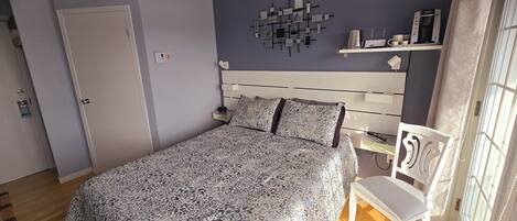 A bedroom with a bed, a nightstand, a chair, a wall-mounted shelf, and a window with curtains.
