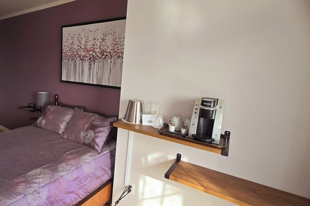 Un dormitorio con una cama, una mesita de noche con lámpara, una cafetera y una estantería con diversos objetos.