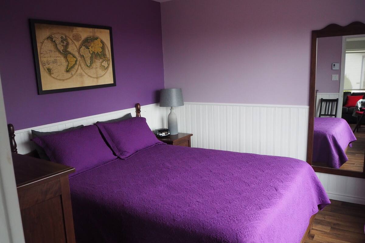 Un dormitorio con una cama grande, ropa de cama morada, un mapa en la pared, una mesita de noche, una lámpara y un espejo.