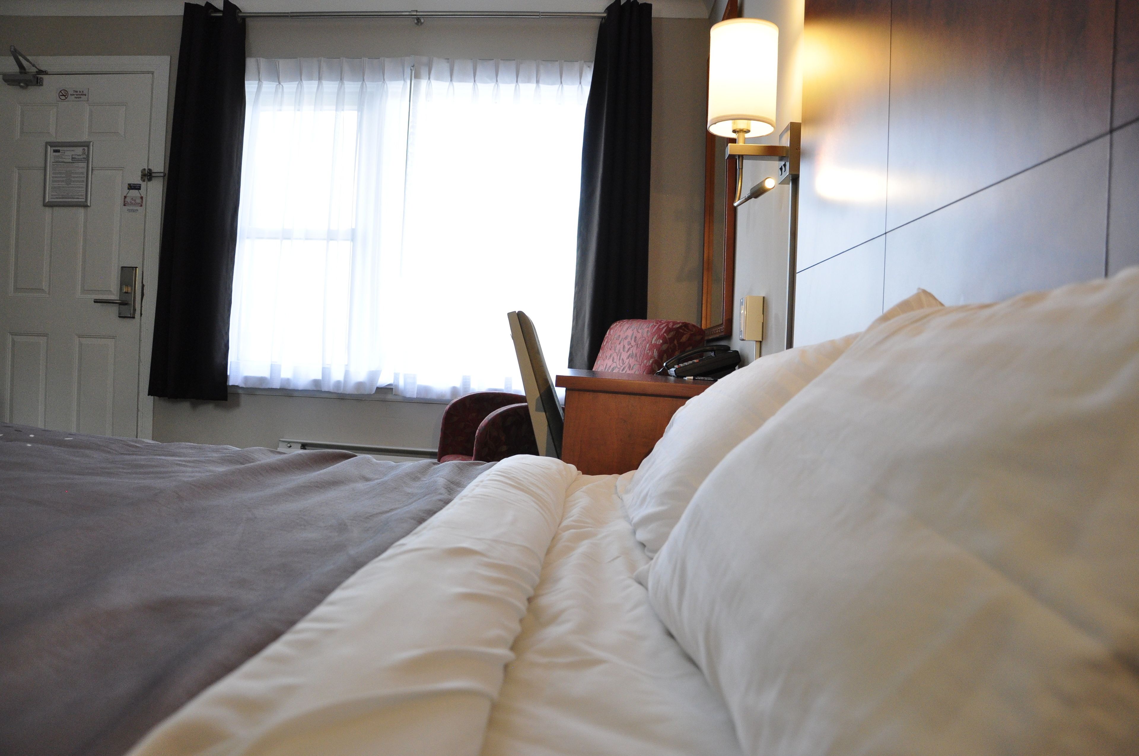 A hotel room with a bed, a chair, a desk with a telephone, a lamp, and a window with curtains.