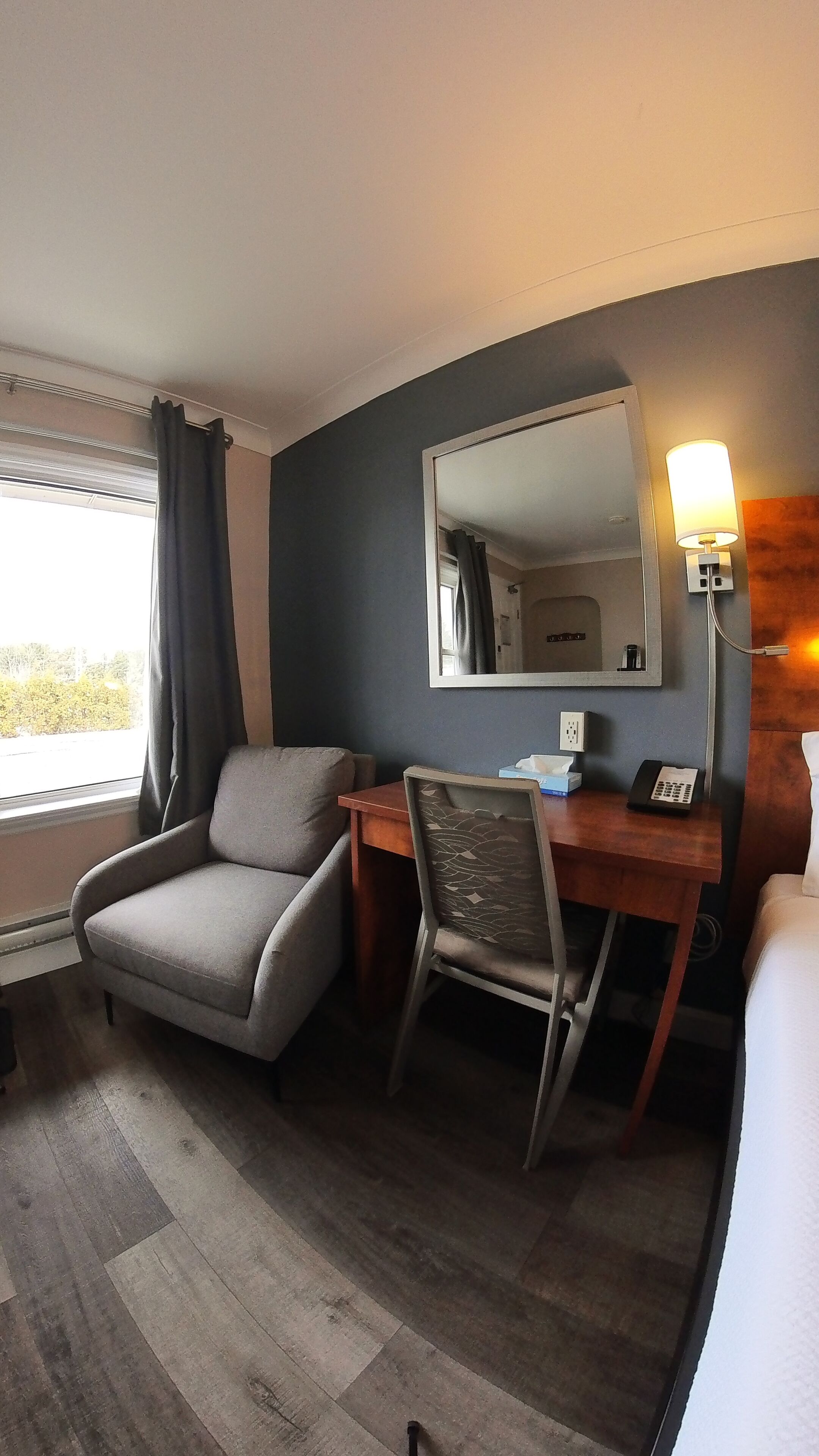A hotel room with a grey armchair, a wooden desk with a telephone, a mirror, and a bedside table with a lamp.