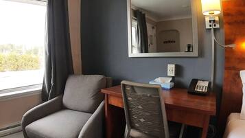 A hotel room with a grey armchair, a wooden desk with a telephone, a mirror, and a bedside table with a lamp.