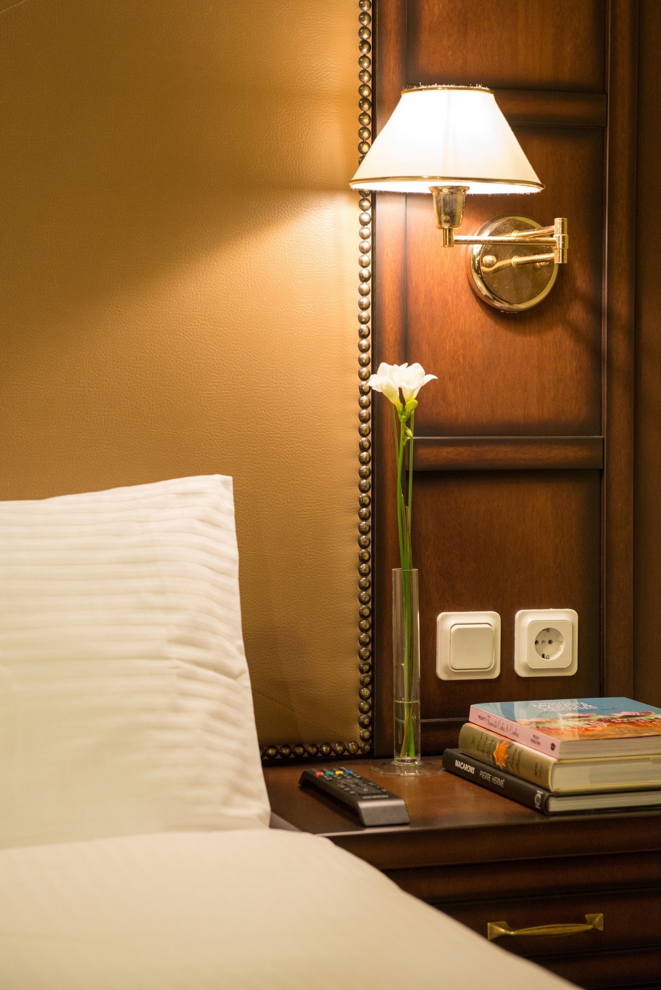 Een hotelkamer met een bed, een nachtkastje met een lamp, een vaas met een bloem en een boek.