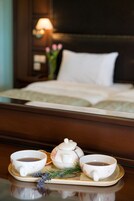 Cama bem arrumada com travesseiro branco e cabeceira escura. Em frente à cama, um suporte com chaleira, duas xícaras e um raminho de lavanda.
