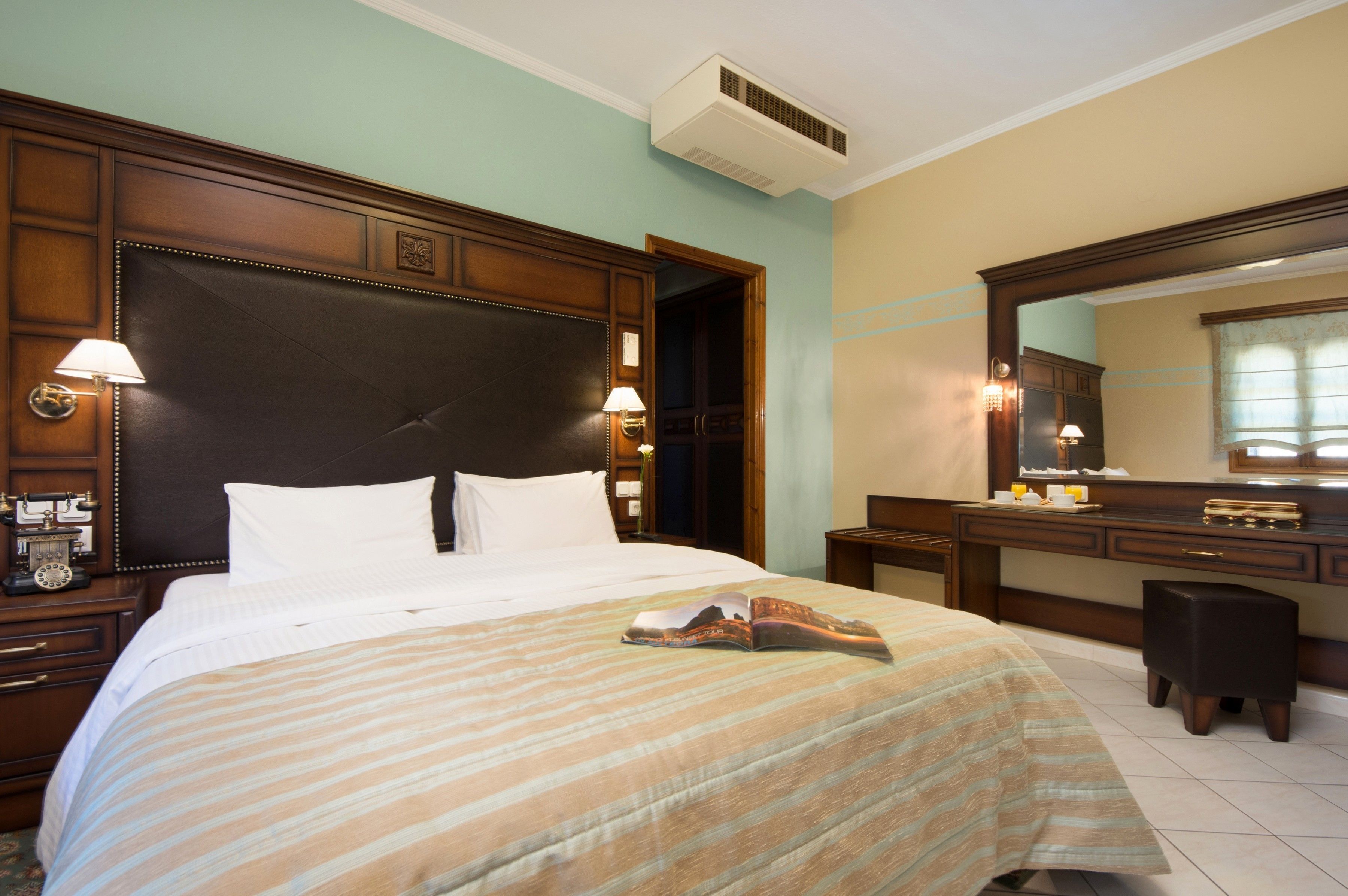 Ein Hotelzimmer mit einem großen Bett, einem Holzkopfteil, einem Nachttisch mit Lampe, einem Schminktisch und einem Spiegel.