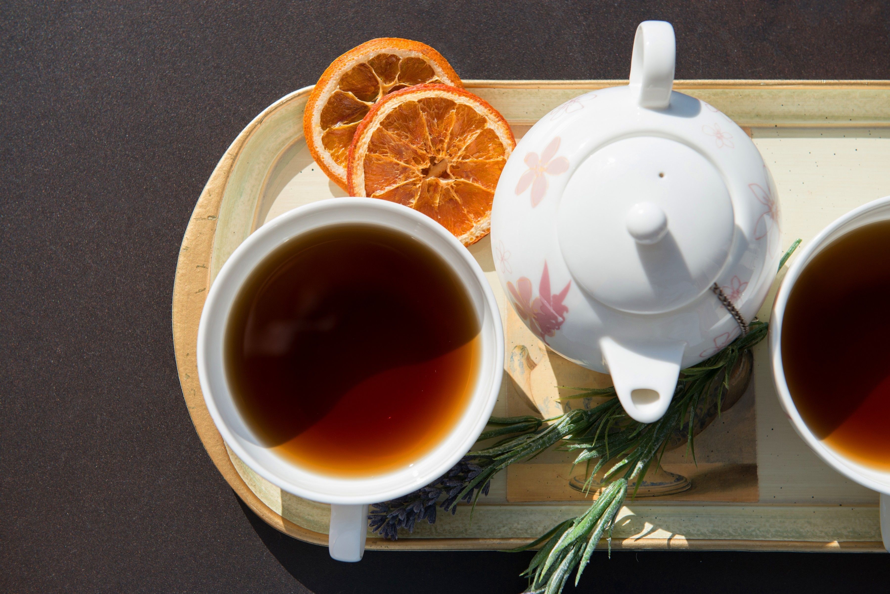 A tray with two teacups and a teapot, accompanied by orange slices and a sprig of rosemary.