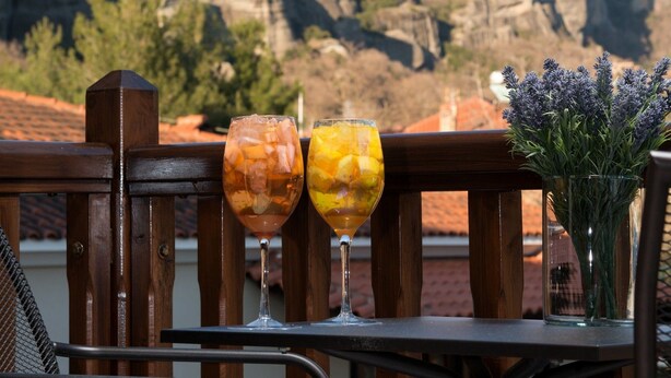 A balcony with a view of rock formations, a wooden railing, and a table with two glasses of drinks and a vase of flowers. - Hotel Kastraki (Kalabaka)