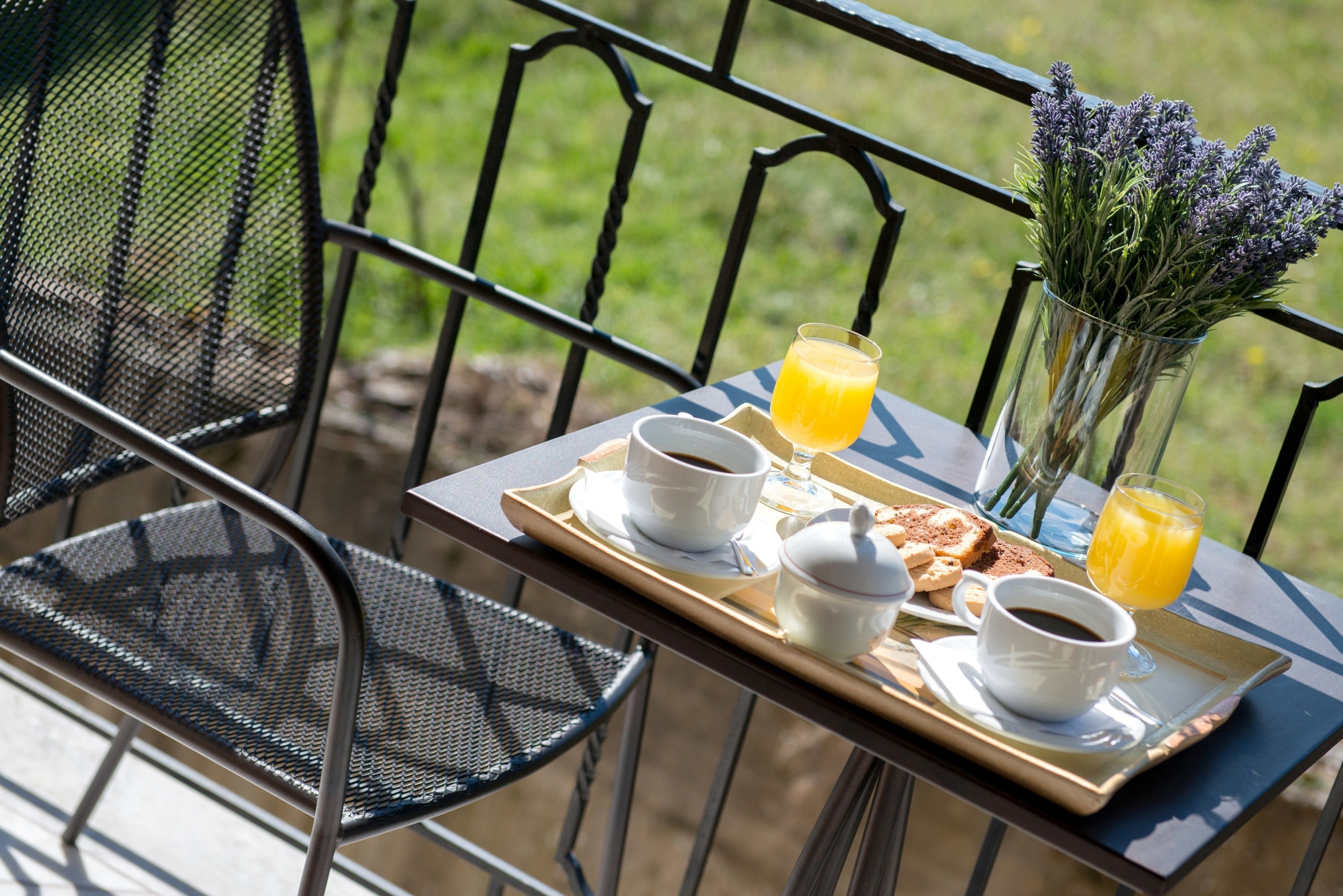 A breakfast tray with coffee, tea, toast, and orange juice on a balcony with chairs and a vase of lavender.