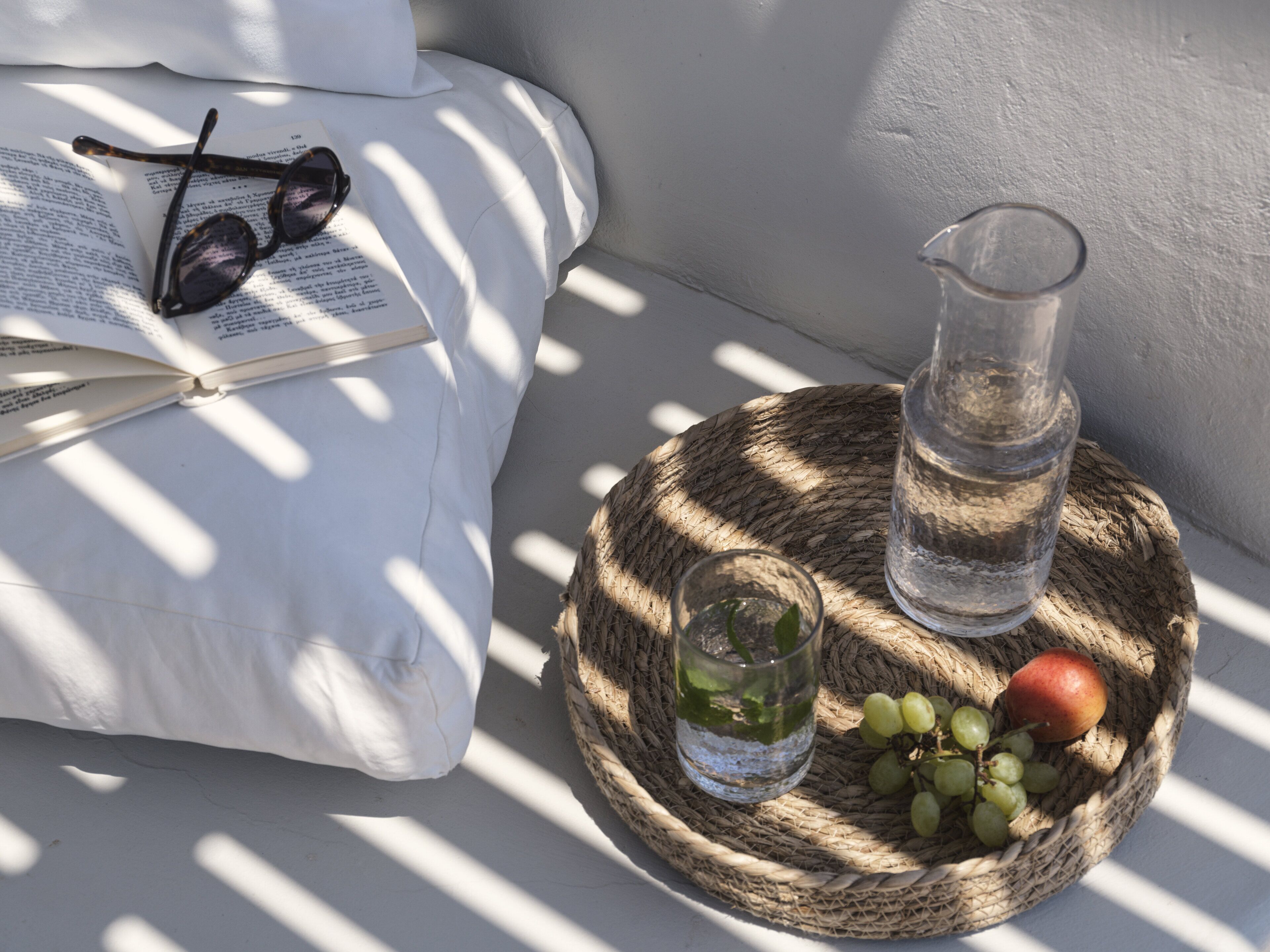 A book, glasses, a glass pitcher, a glass with a straw, and a basket with grapes and an apple on a woven surface.