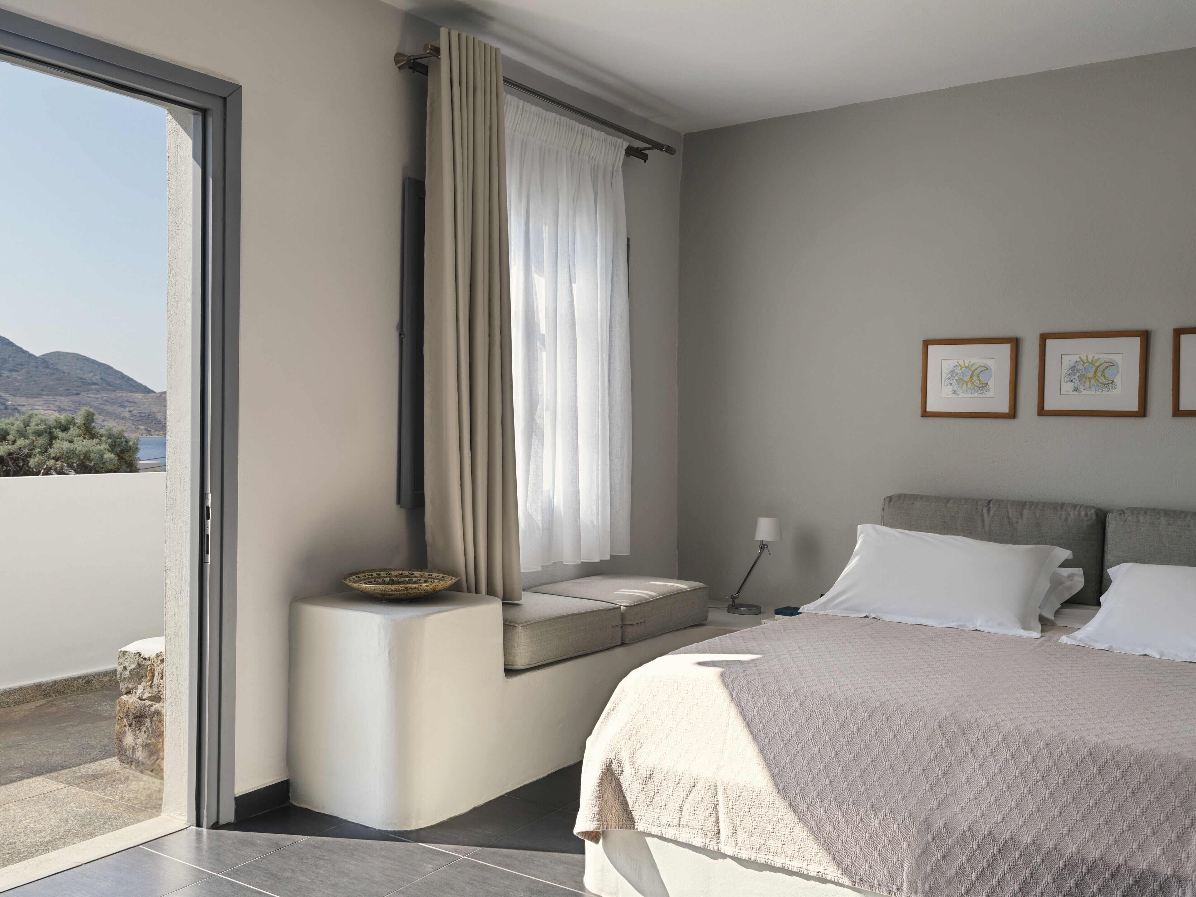 Une chambre à coucher moderne avec un grand lit, une table de chevet, une fenêtre avec des rideaux et des œuvres d’art encadrées accrochées au mur.