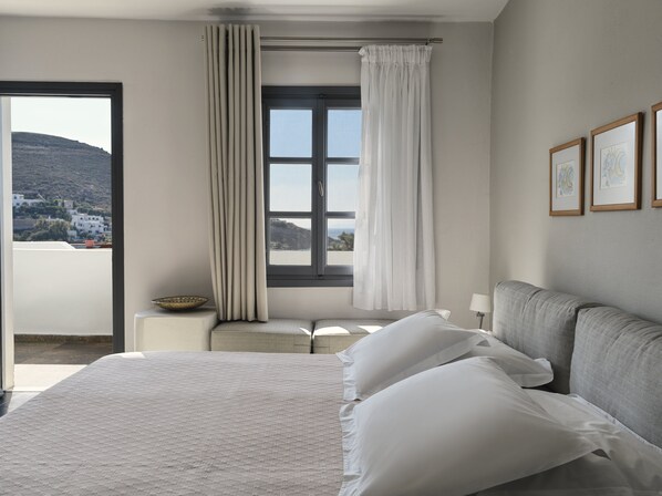 A hotel room with a large bed, a view of a mountainous landscape, a window with curtains, and framed artwork on the wall. - The_Petra, Small Luxury Hotels of the World (Patmos)