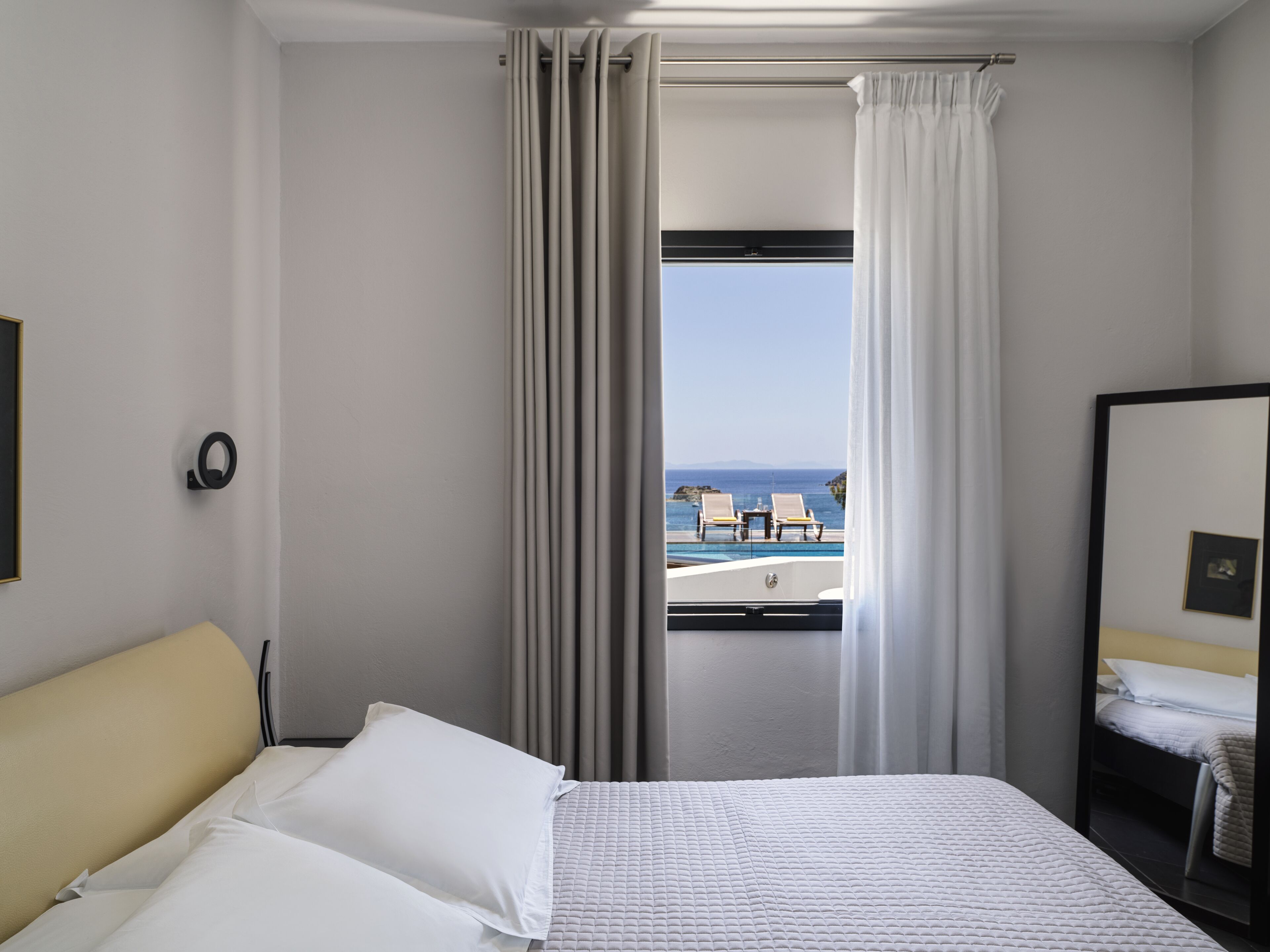 Habitación de hotel con cama, un ventanal con cortinas, vista al mar y una pequeña mesa con sillas afuera.