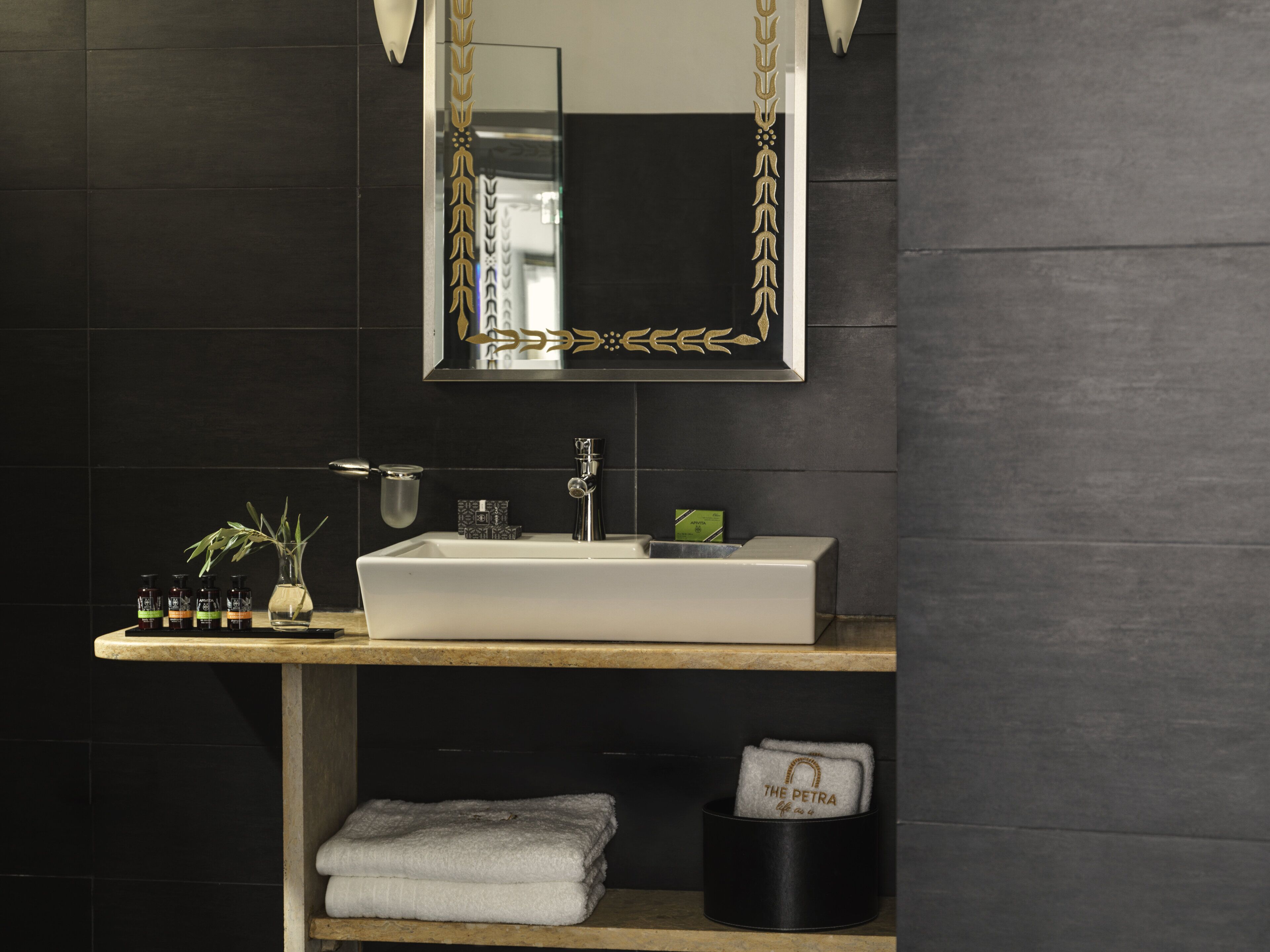 Un baño moderno con un lavabo blanco, baldosas oscuras, un espejo con marco decorativo, una planta pequeña y un toallero.