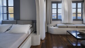 Een moderne slaapkamer met een bed, een zithoek met kussens en uitzicht op zee door grote ramen.