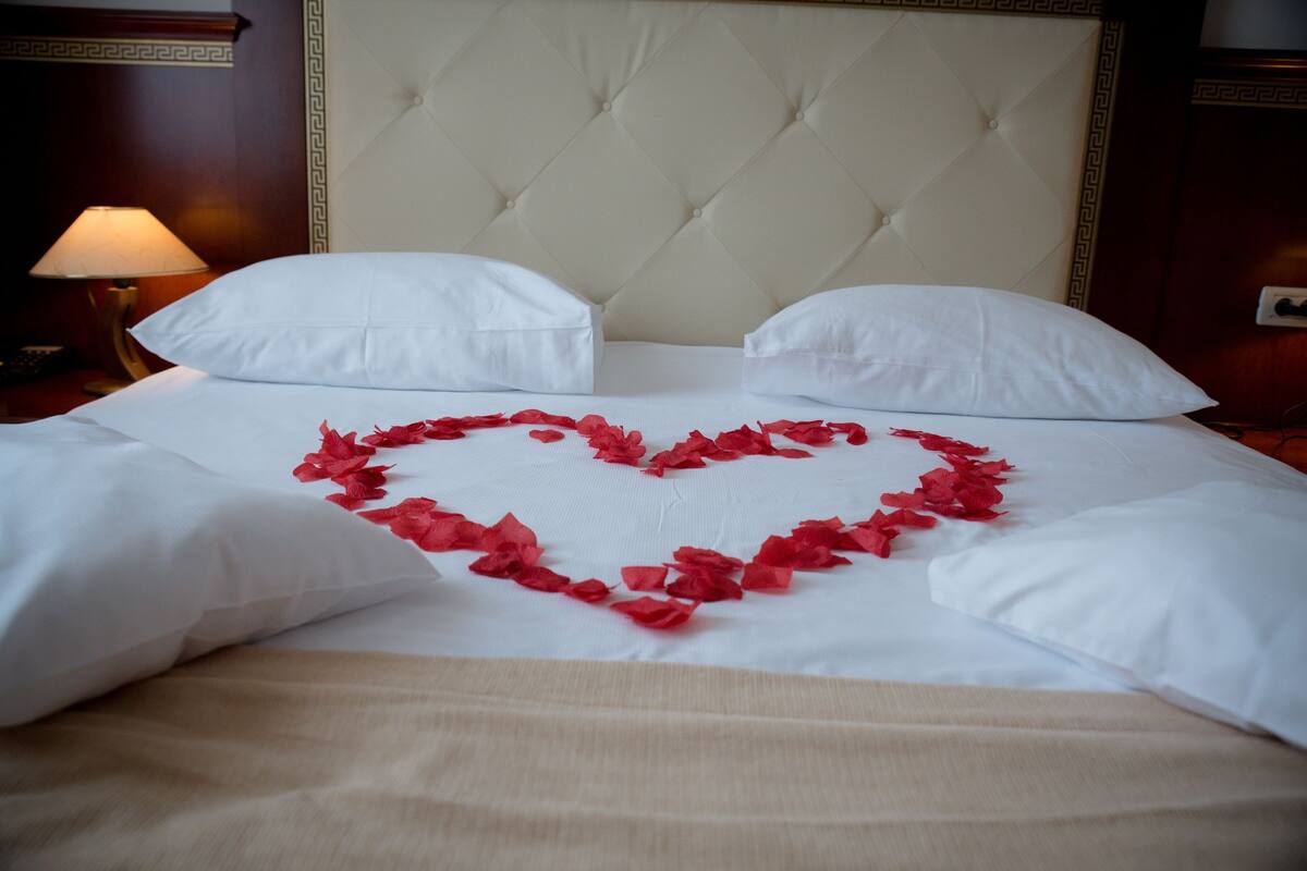 A bed with white linens and a heart-shaped arrangement of red rose petals.