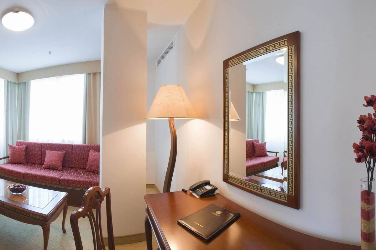 A hotel room with a bed, a desk with a computer, a red sofa, a small table, and a mirror.