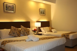 Ein Hotelzimmer mit zwei Betten, jeweils mit weißer Bettwäsche und gemusterten Kissen. An den Wänden hängen Bilder in Rahmen und auf dem Nachttisch steht eine Lampe.