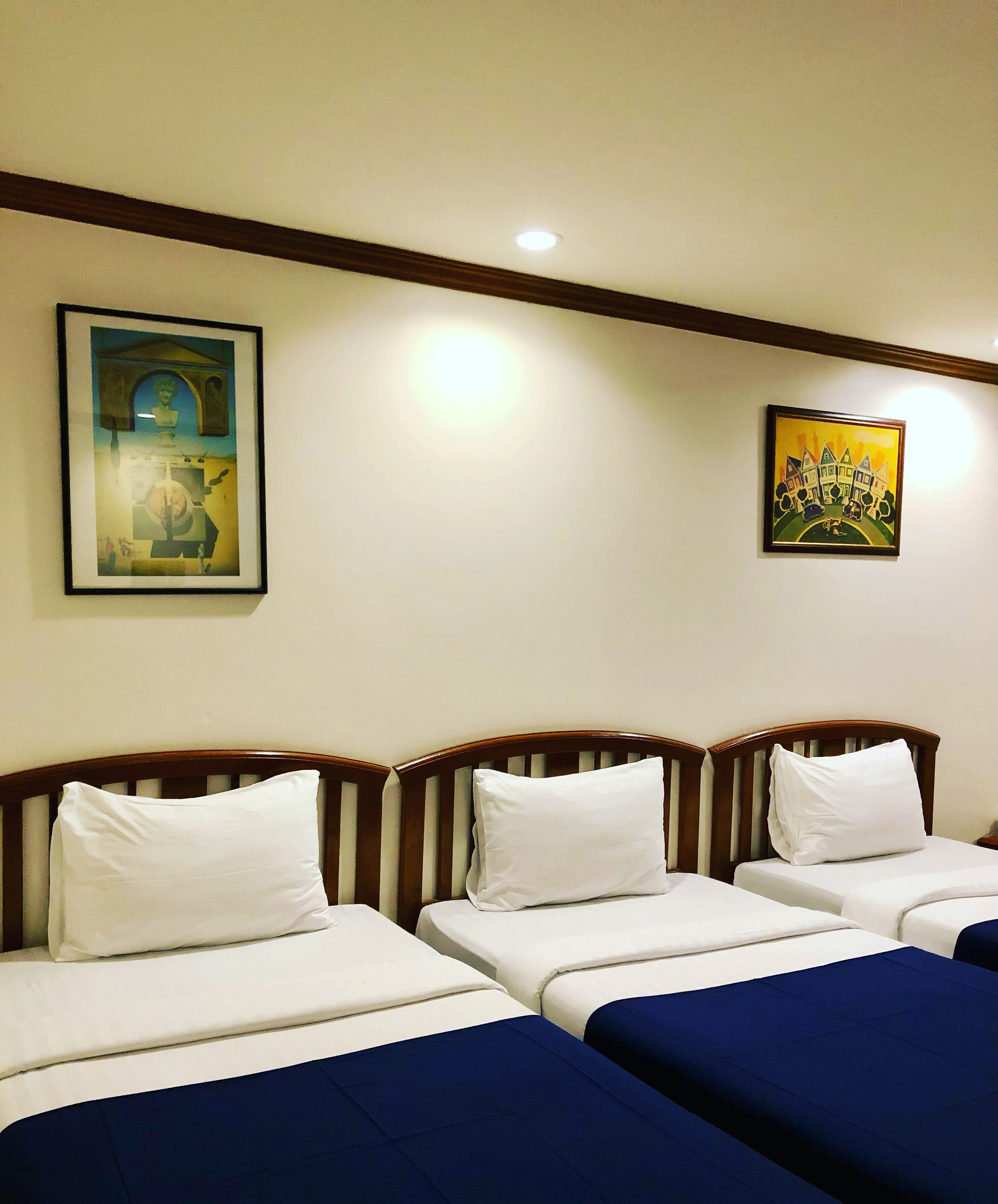A hotel room with three beds, each with white bedding and blue throw pillows. There are two framed artworks on the wall.