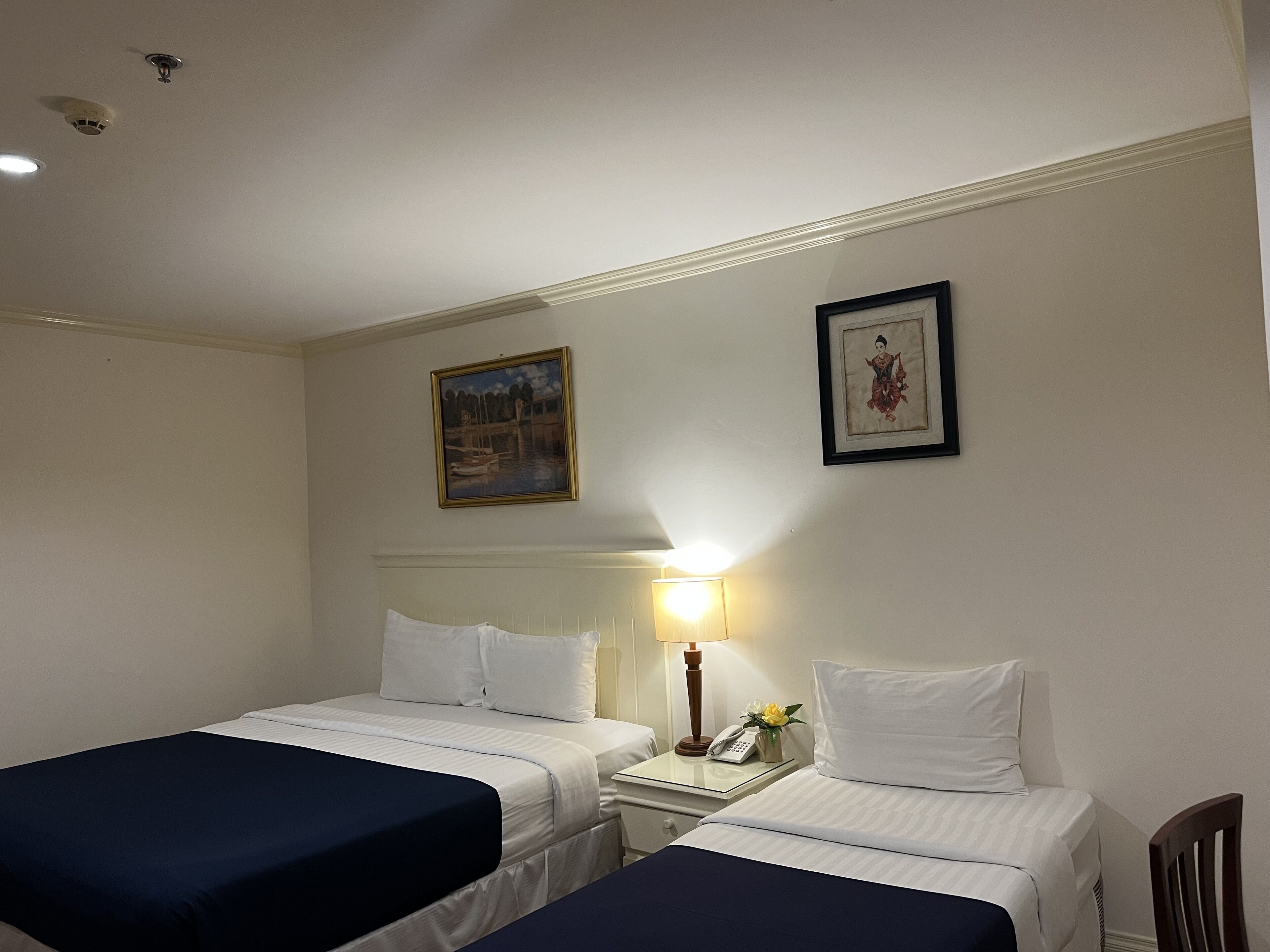 A hotel room with two single beds, a nightstand with a lamp, a framed picture on the wall, and a small table with a vase of flowers.