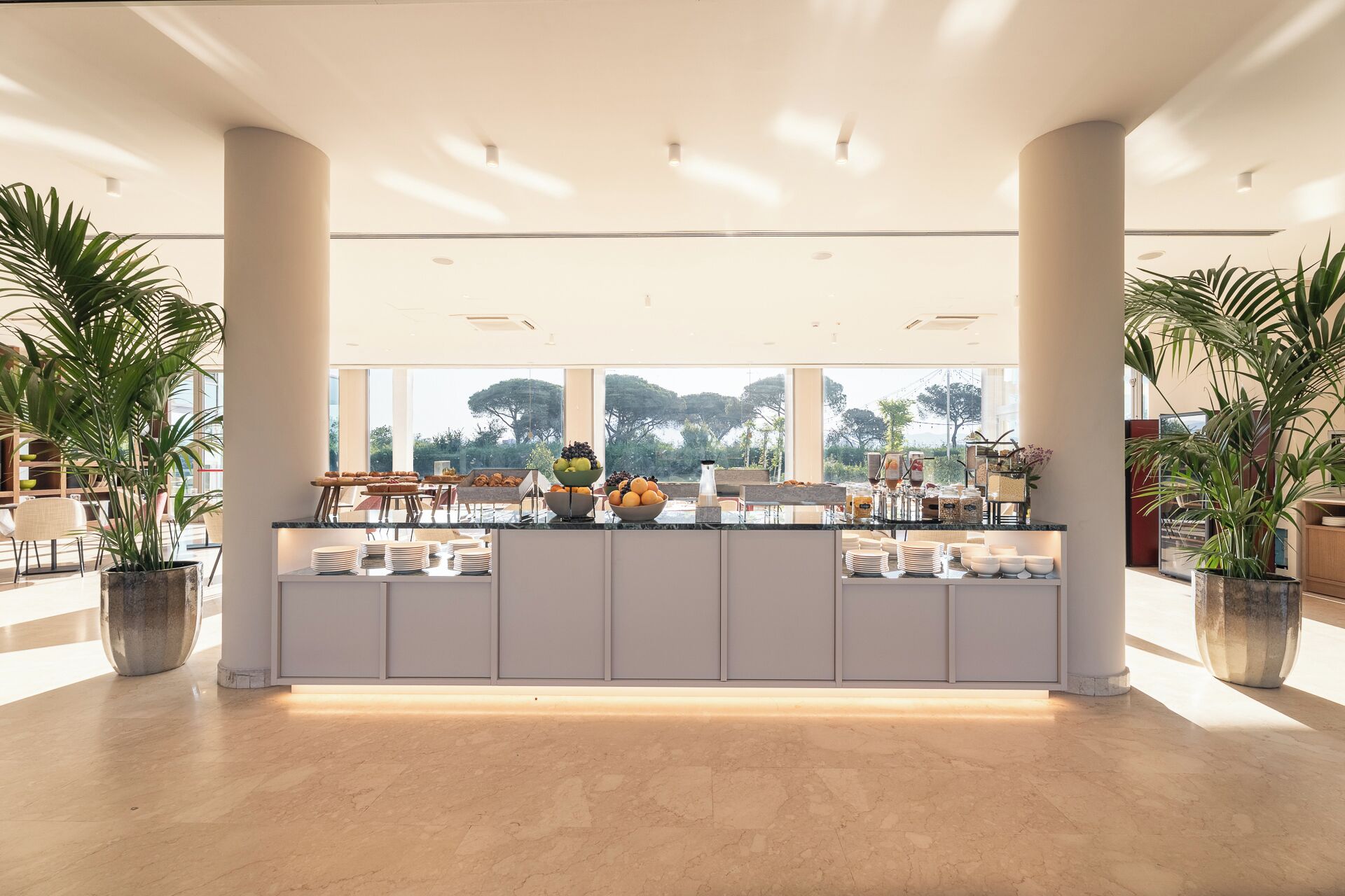 Eine geräumige Hotelhalle mit einem Marmor-Rezeptionstresen, großen Topfpflanzen und Blick ins Freie.