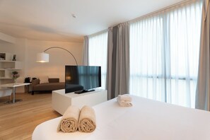 Una habitación de hotel moderna con televisión de pantalla plana, un sofá, un escritorio con lámpara y un amplio ventanal con cortinas.