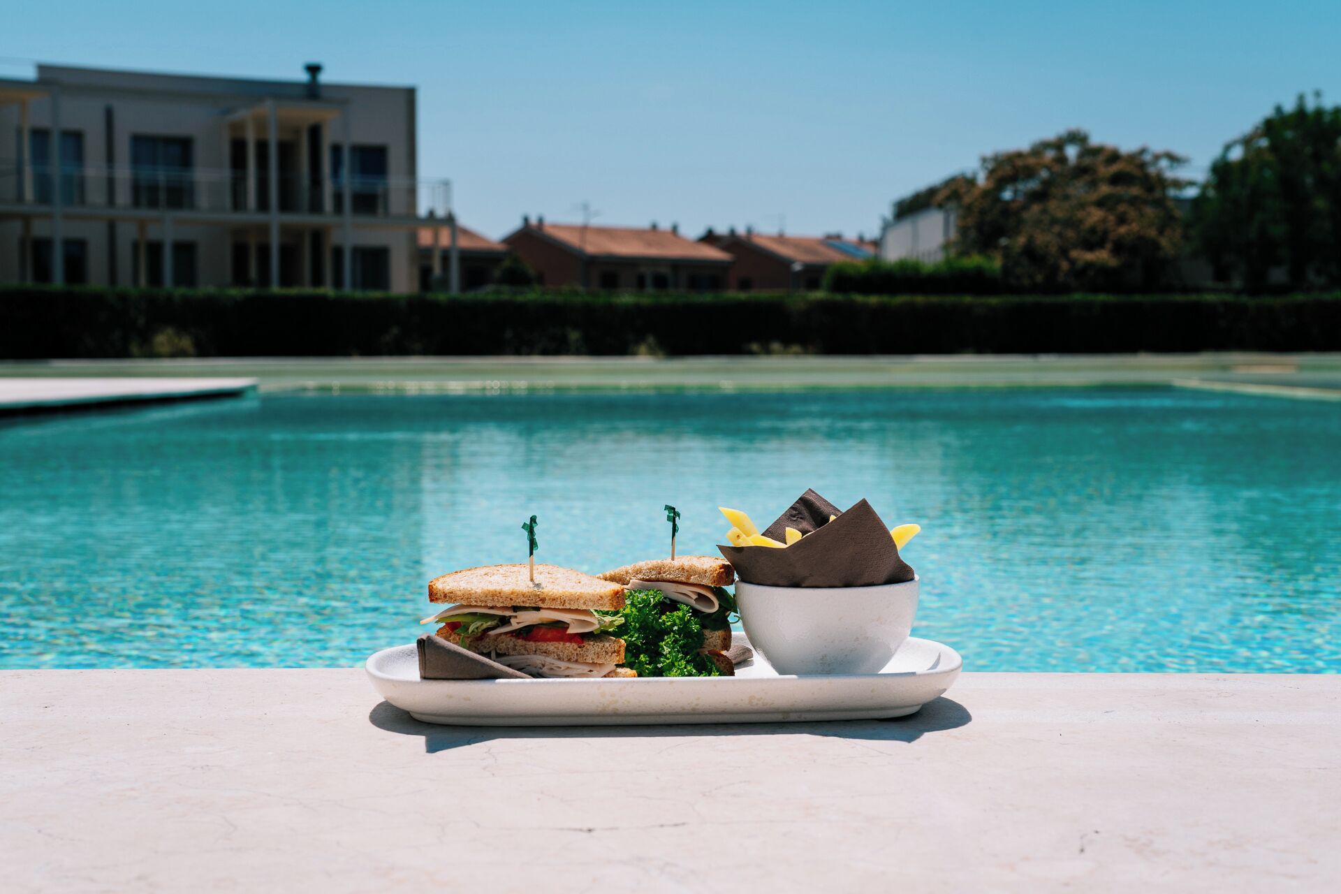 A dining table with a plate of sandwiches and a bowl of fruit by a poolside.