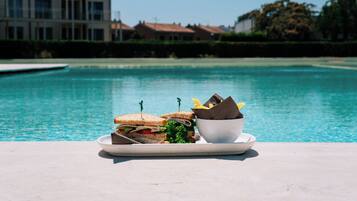 A dining table with a plate of sandwiches and a bowl of fruit by a poolside.