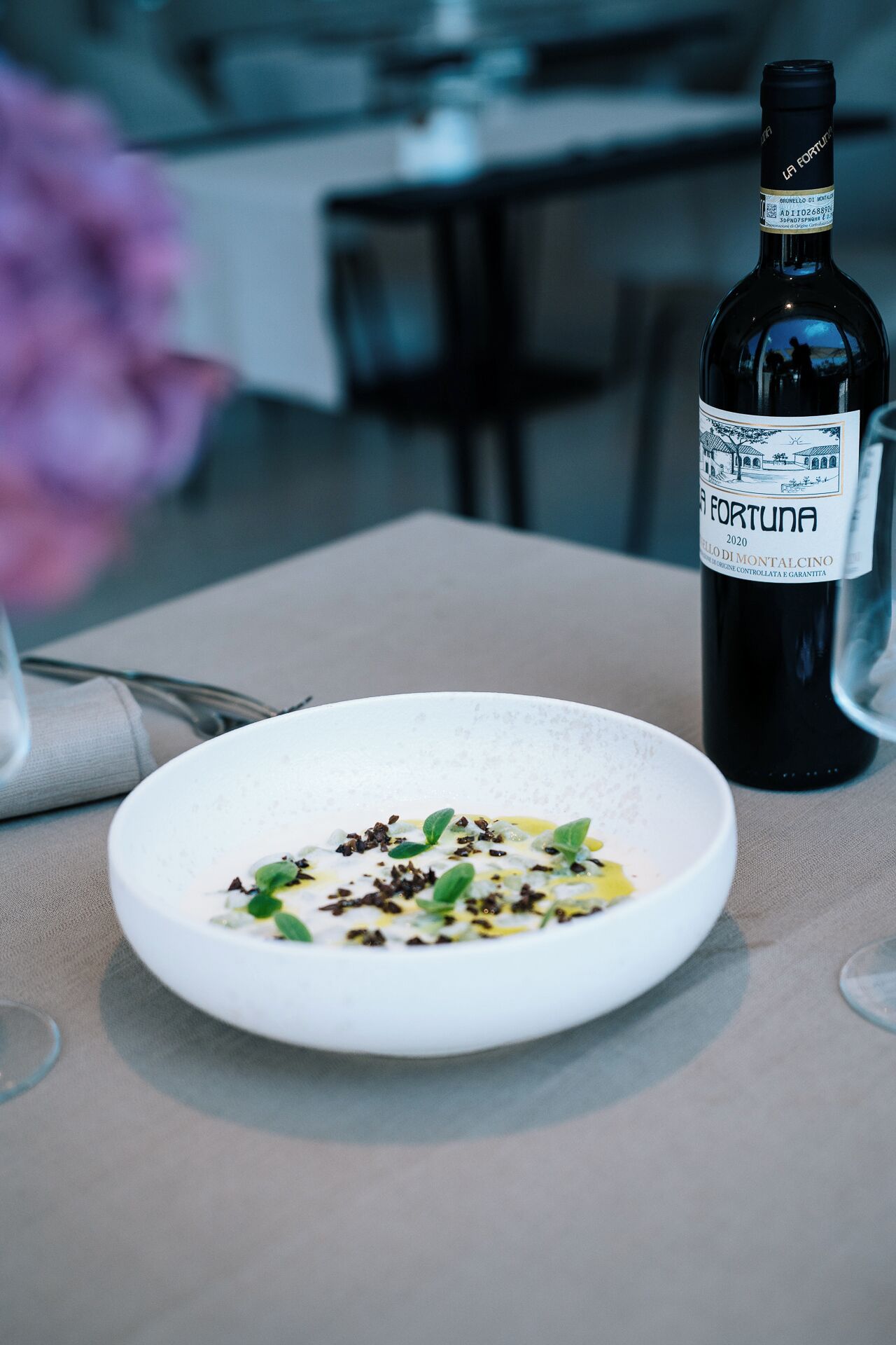 Una mesa de comedor con un plato de comida blanca, una botella de vino y un jarrón con flores.