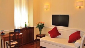 Habitación de hotel con sofá blanco, cojines rojos, televisor de pantalla plana en la pared, una mesita con un florero y una mesa de comedor de madera con sillas.