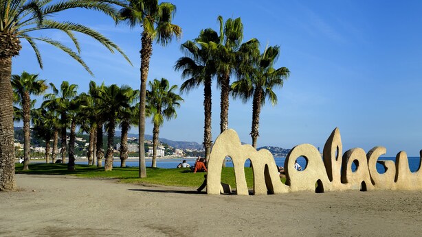 Una playa con palmeras y una gran escultura que deletrea 'Málaga' sobre la arena.