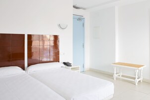 A hotel room with two beds, a wooden headboard, a small bedside table with a phone, a light blue door, and a white bench.