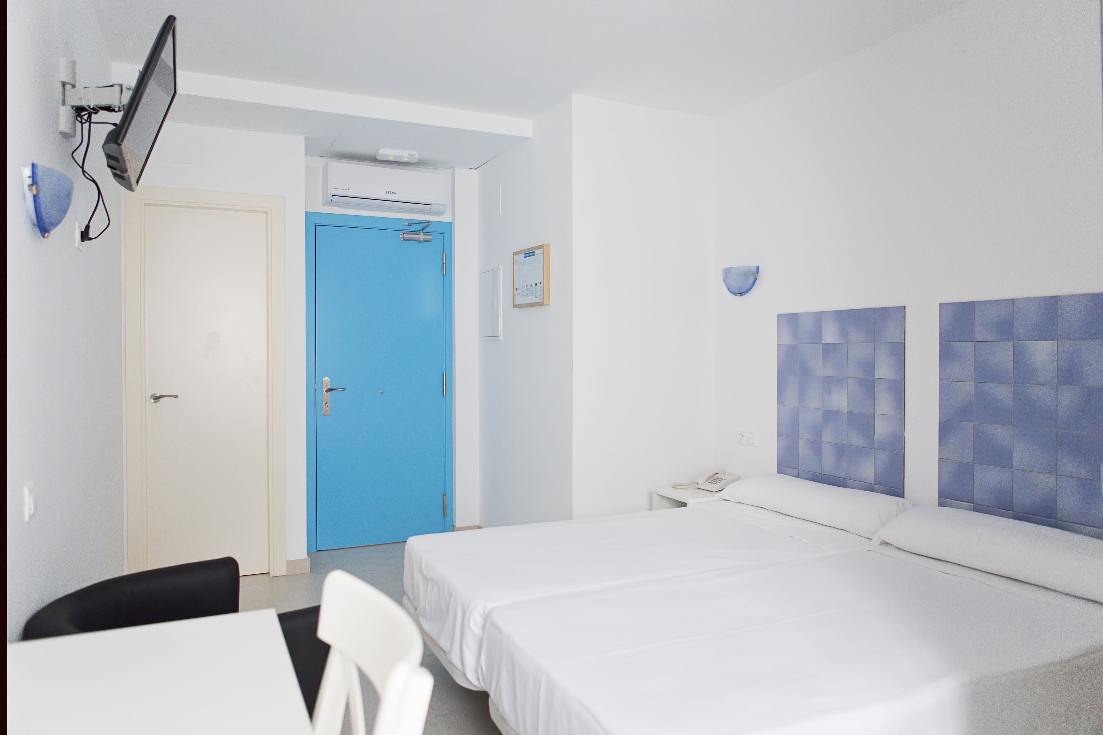 A hotel room with a bed, a desk, a chair, a blue door, and a wall-mounted phone.