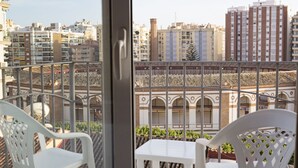 Un balcón con sillas blancas y una mesa, con vistas a un paisaje urbano que incluye edificios y una estructura de tejados con tejas.