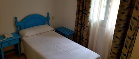 Una cama individual con cabecera azul, una mesita de noche con un teléfono y una ventana con cortinas.