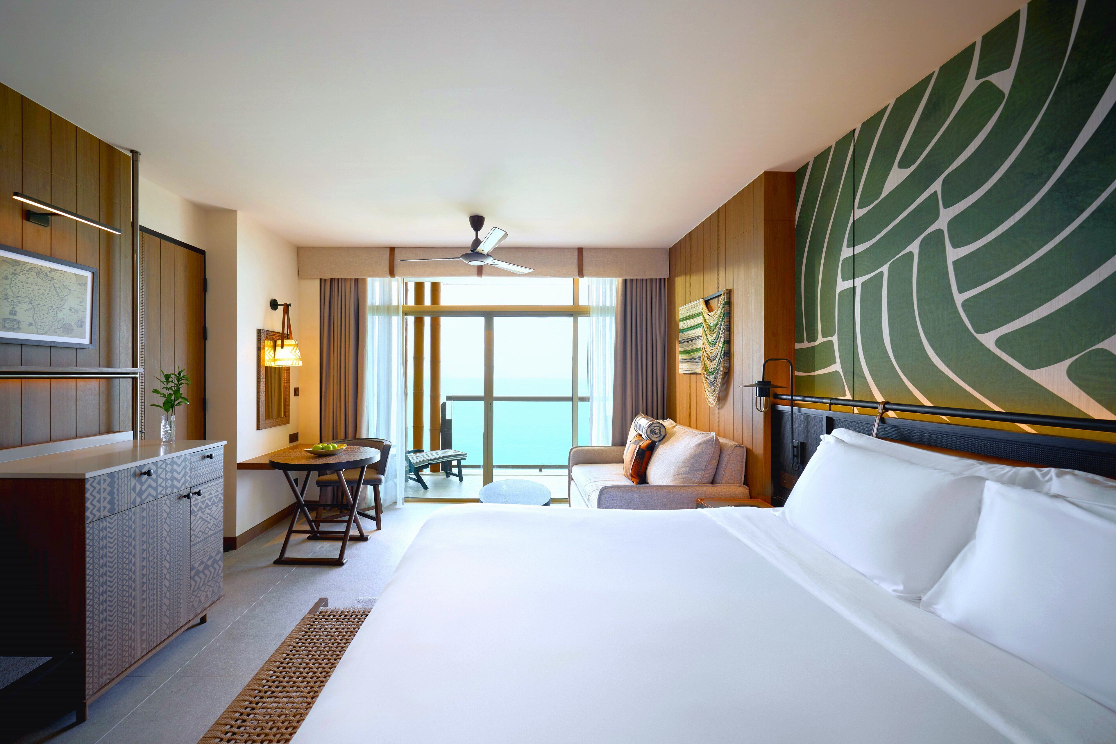 A hotel room with a large bed, a desk with a chair, a small table, a sofa, and a view of the sea through large windows.