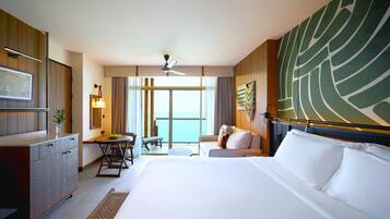A hotel room with a large bed, a desk with a chair, a small table, a sofa, and a view of the sea through large windows.