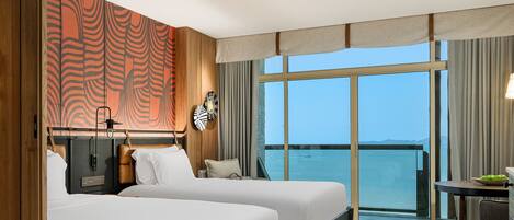 A hotel room with two beds, a large window with a view of the sea, a ceiling fan, and a small table with a chair.