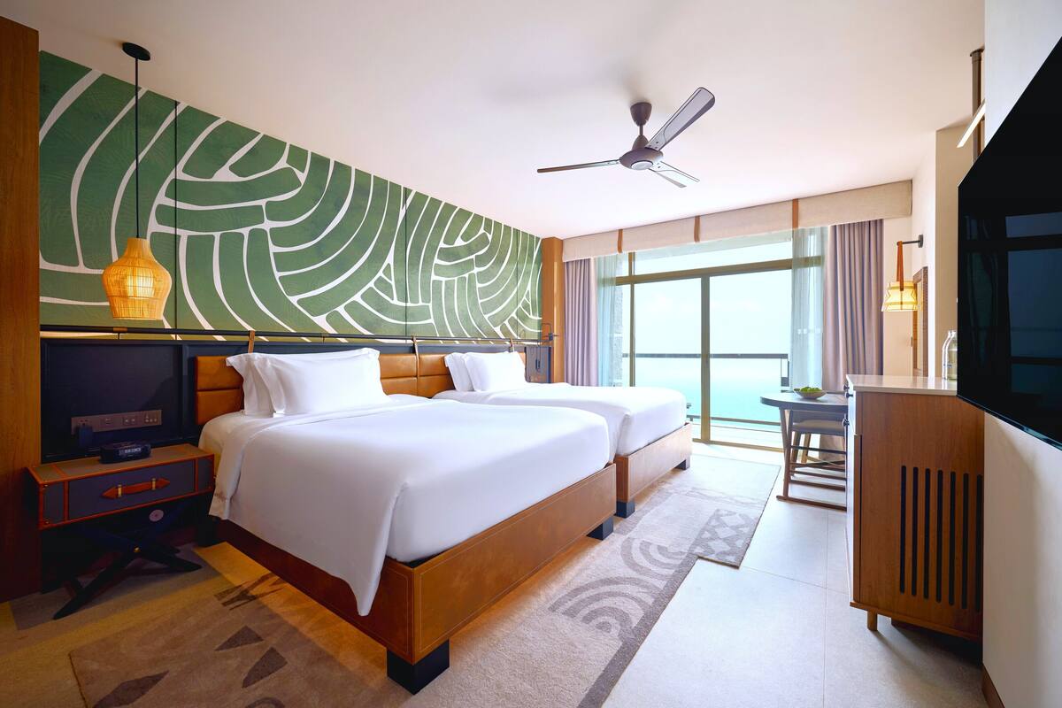 Habitación de hotel con dos camas, televisión, escritorio y un amplio ventanal con cortinas.