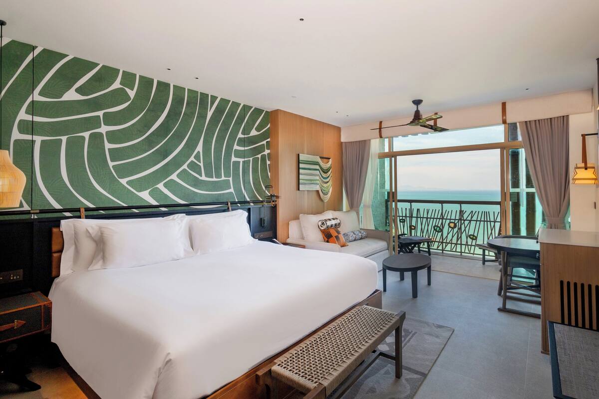 Habitación de hotel con una cama grande, un sofá, una mesita, un balcón con barandilla y vista al mar.