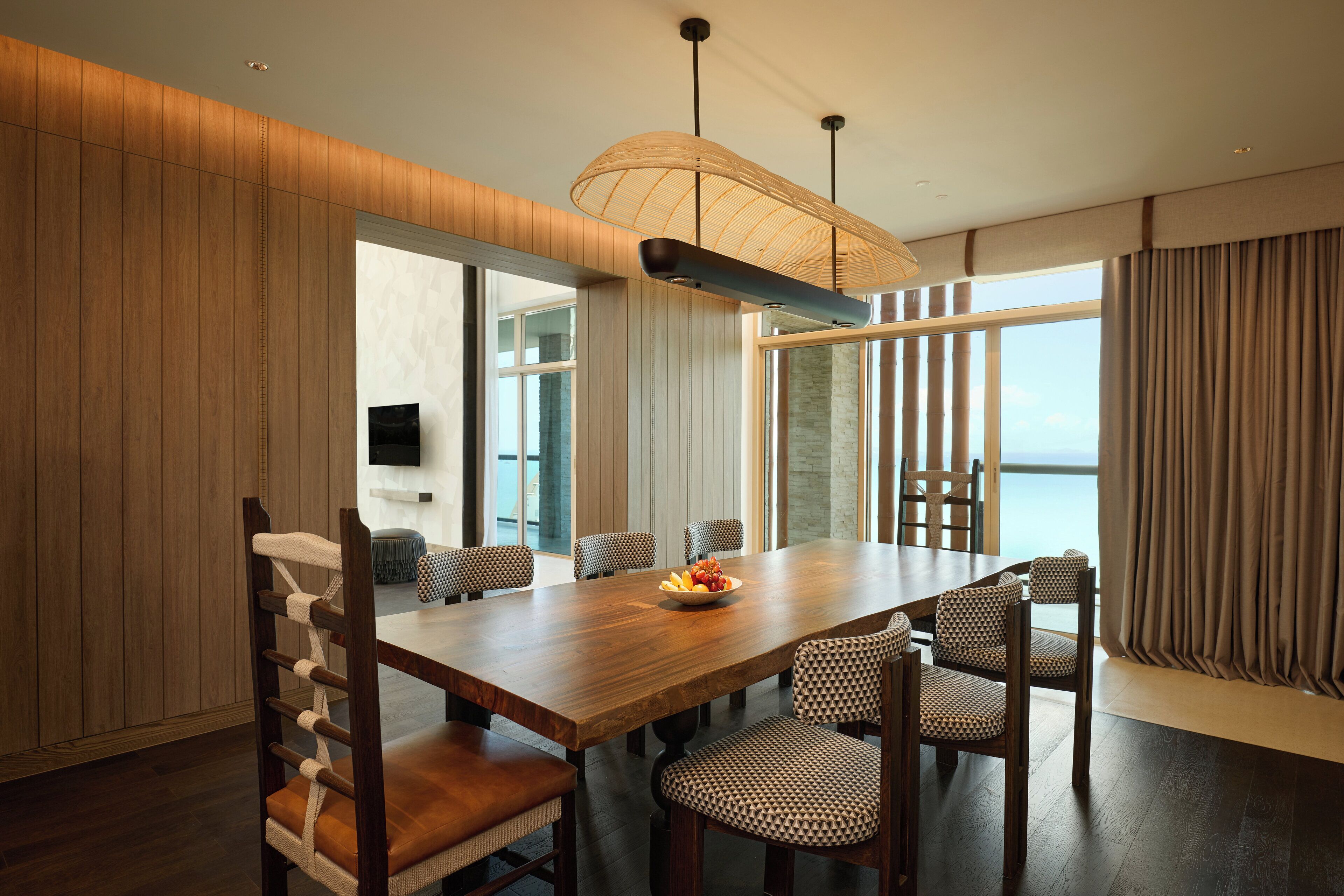 A modern dining area with a wooden table and chairs, a large window with curtains, and a hanging light fixture.