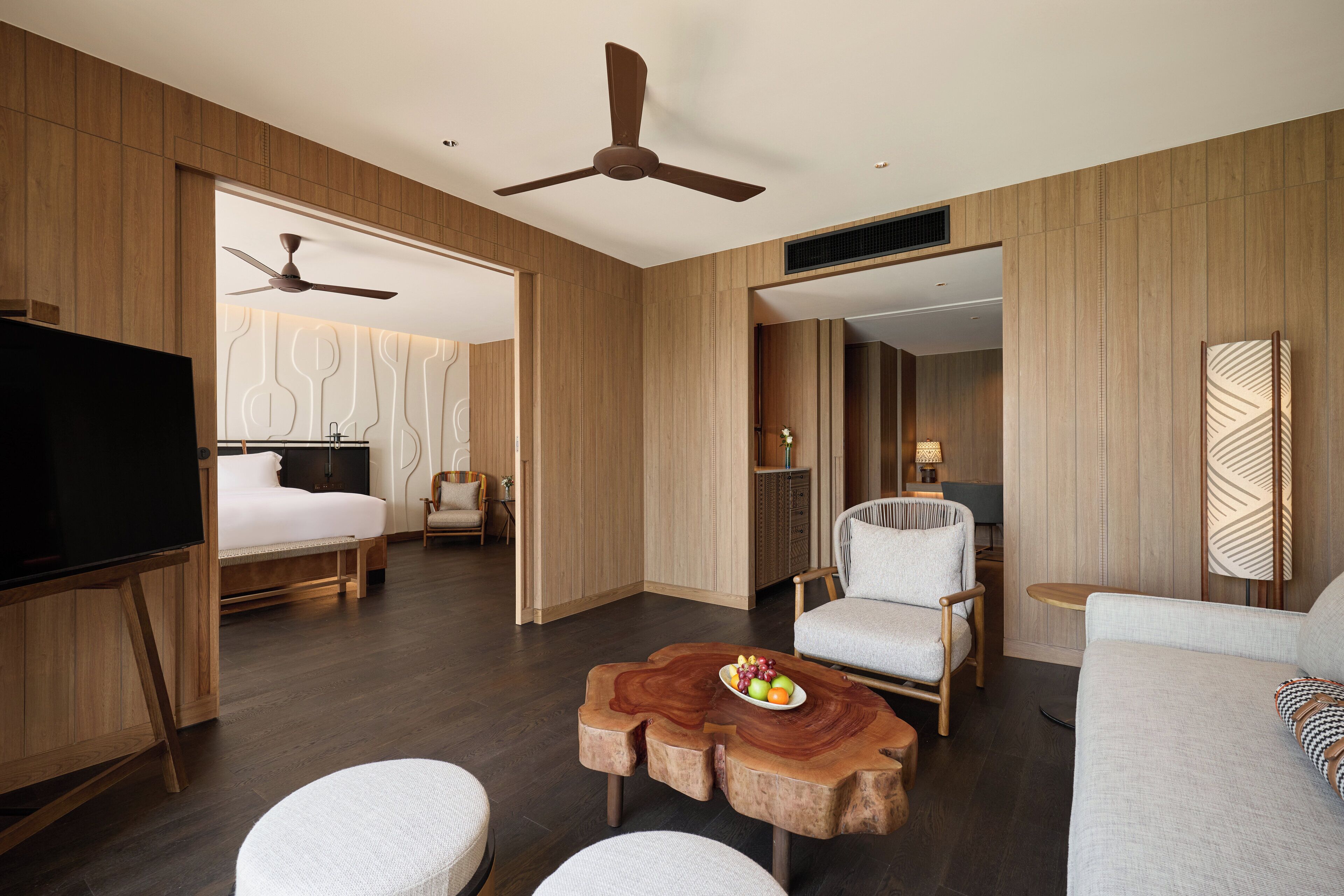 A modern hotel room with a bed, a TV, a ceiling fan, a wooden coffee table with fruit, and a sofa.