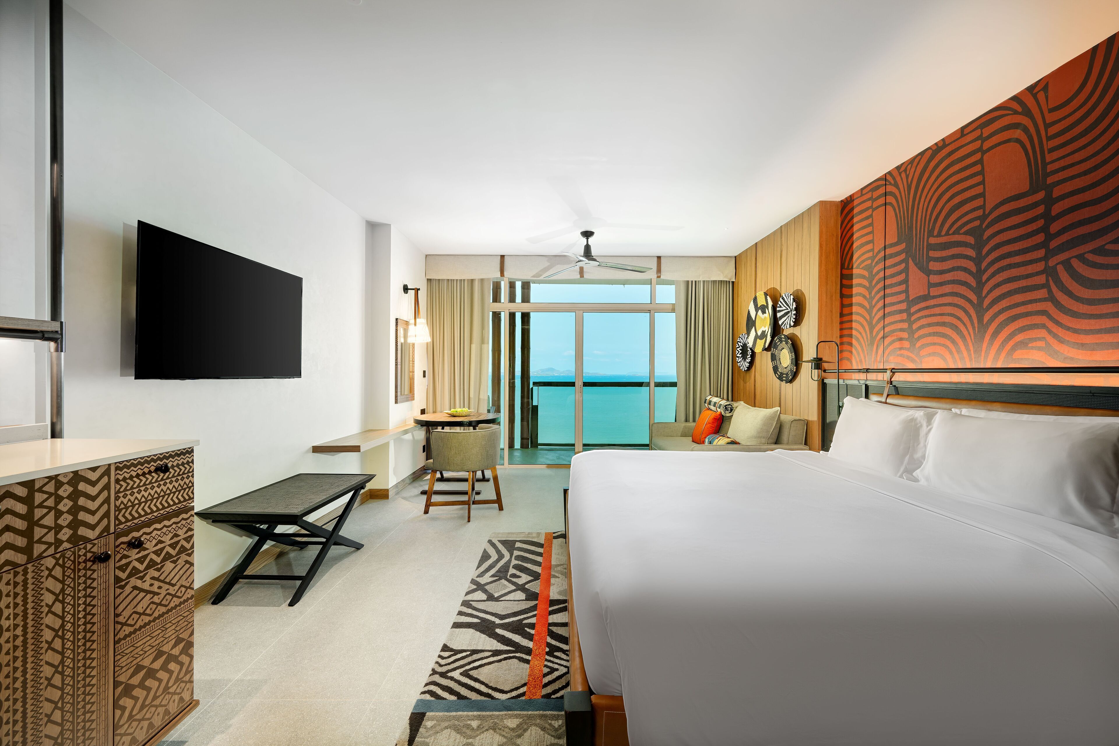 A modern hotel room with a large bed, a desk, a chair, a flat-screen TV, and a balcony with a view of the sea.
