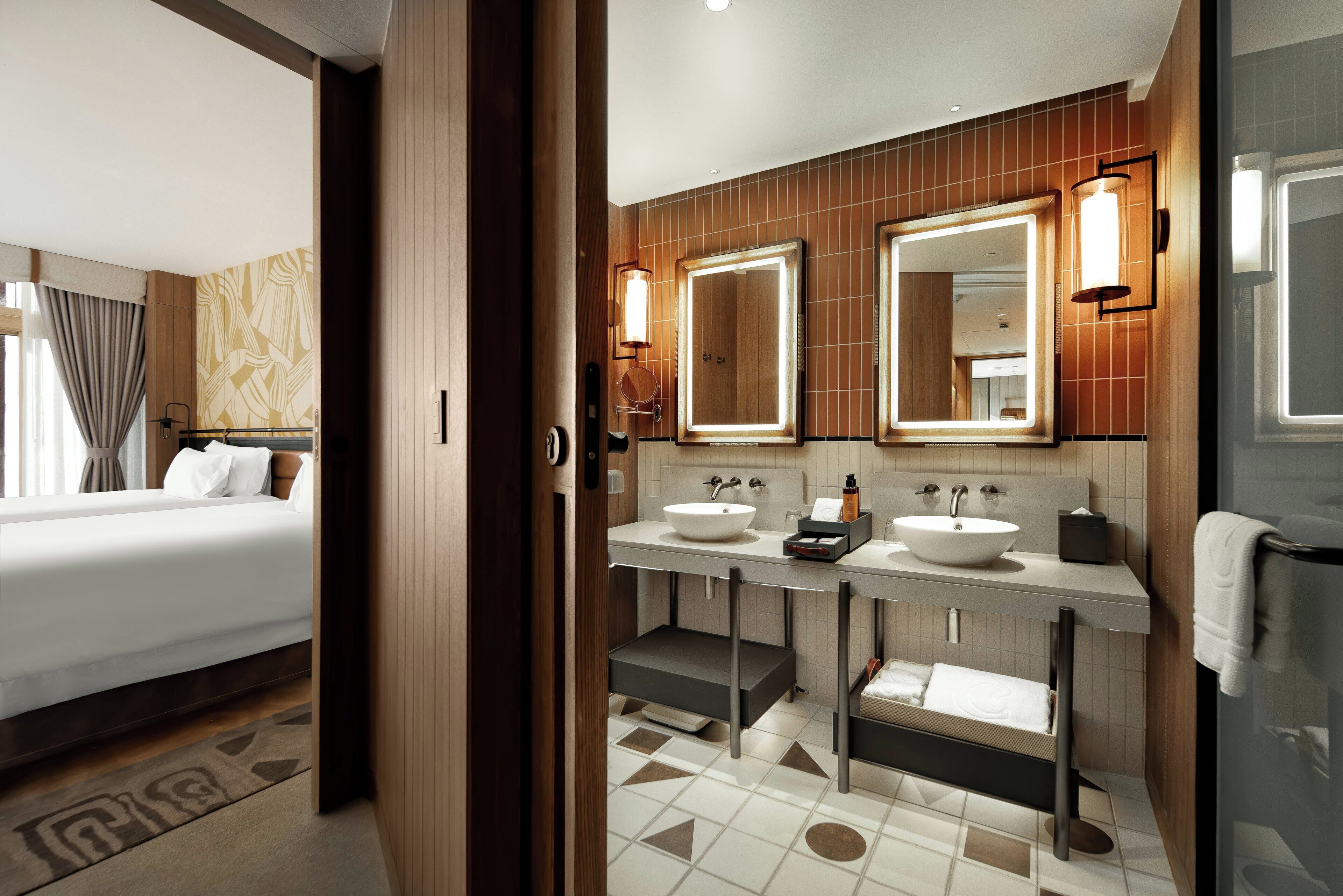Habitación de hotel con cama, baño con dos lavamanos, espejo y ducha.