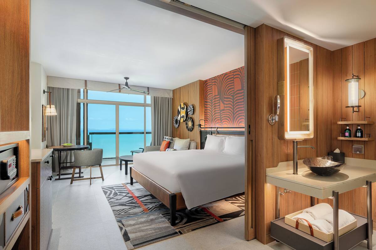 Habitación de hotel con cama, escritorio, silla, baño con lavamanos y espejo, y vista al océano.