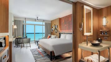 Habitación de hotel con cama, escritorio, silla, baño con lavamanos y espejo, y vista al océano.