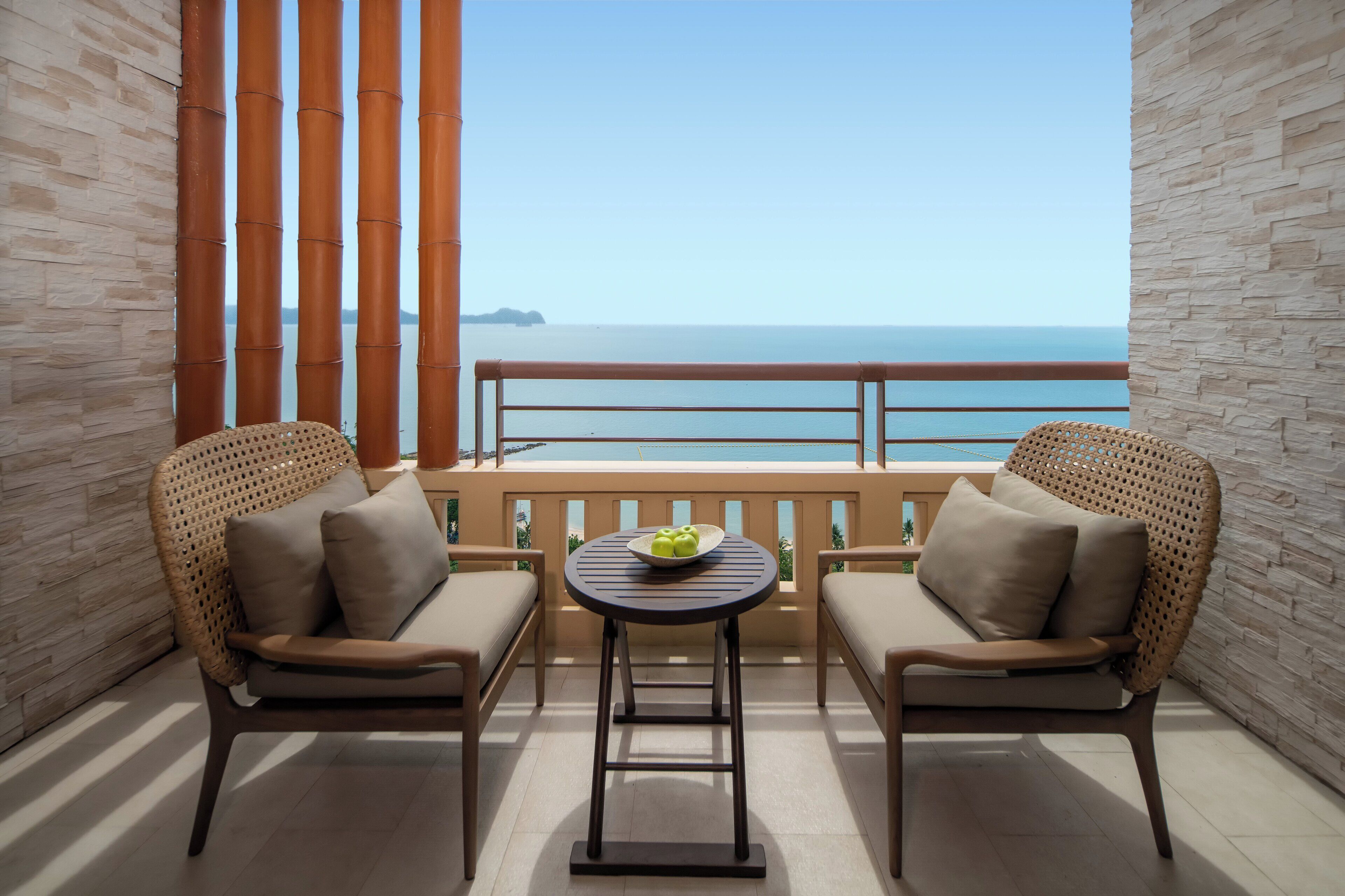 A balcony with two wicker chairs and a small table, featuring a clear view of the sea and distant islands.